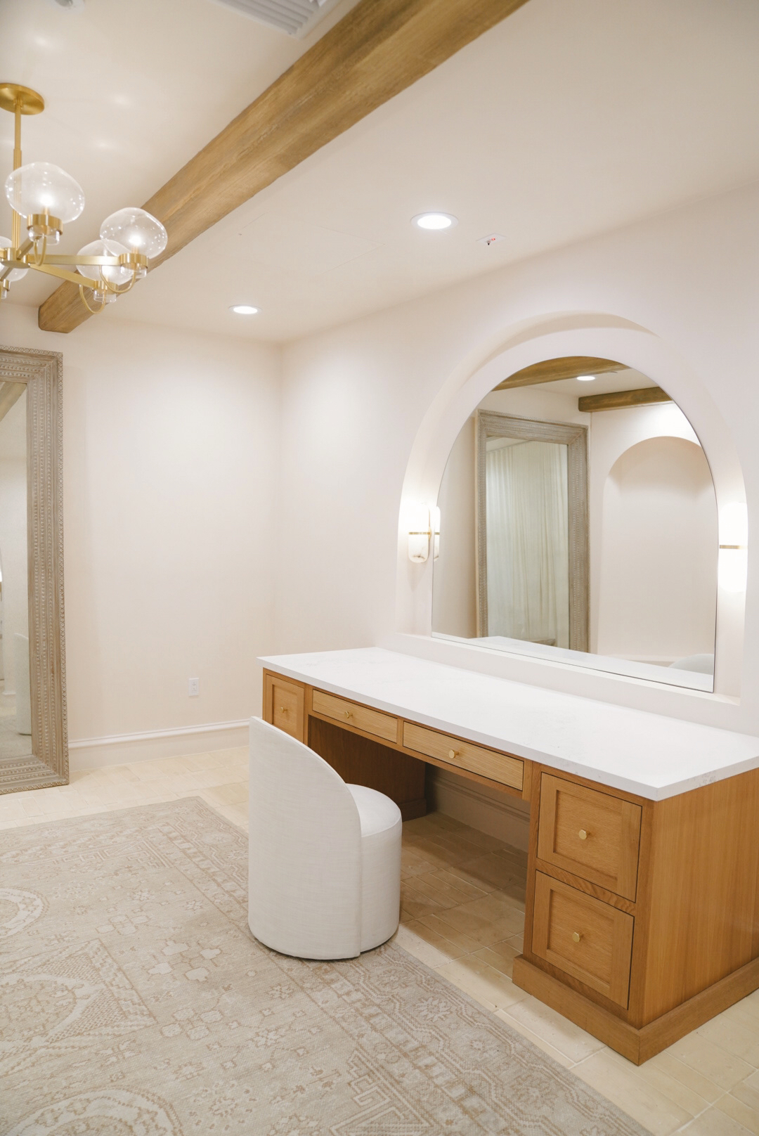 Minimalist vanity with a white marble countertop, wooden drawers, arched mirror, and a round white cushioned stool on a patterned beige rug.