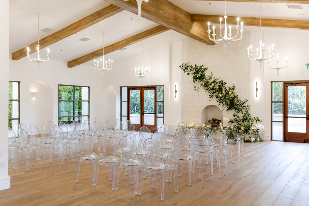 Bright event room with wooden beams, clear ghost chairs arranged in rows, white chandeliers, and a fireplace decorated with green foliage.