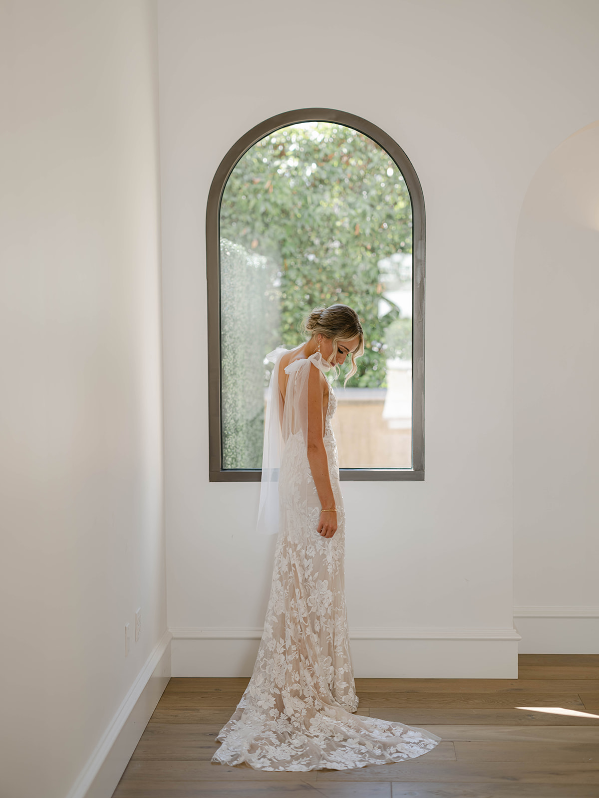 Bride in a white floral lace wedding dress with a long train standing in front of an arched window with greenery outside.