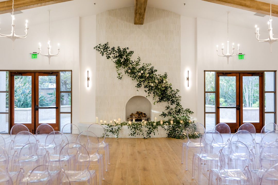 Bright event space with transparent chairs facing a white fireplace decorated with green garlands and candles, flanked by wooden double doors and white chandeliers.