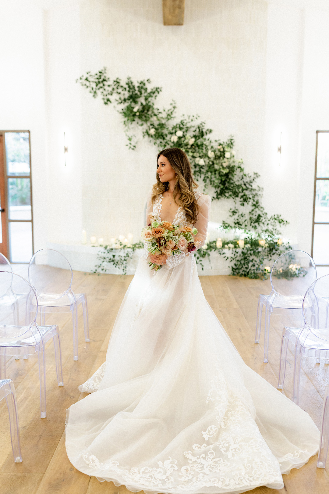 Bride in a long white lace wedding gown holding a bouquet of flowers, standing in a bright room with transparent chairs and greenery decoration on the wall.