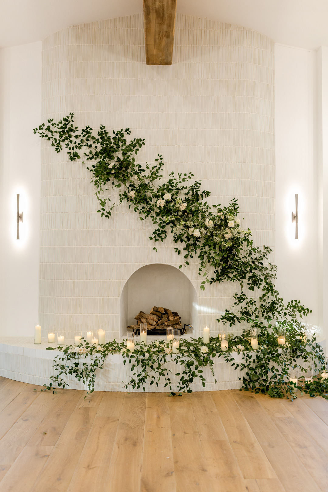 White fireplace decorated with cascading green vines, white flowers, and lit candles on a light wooden floor.
