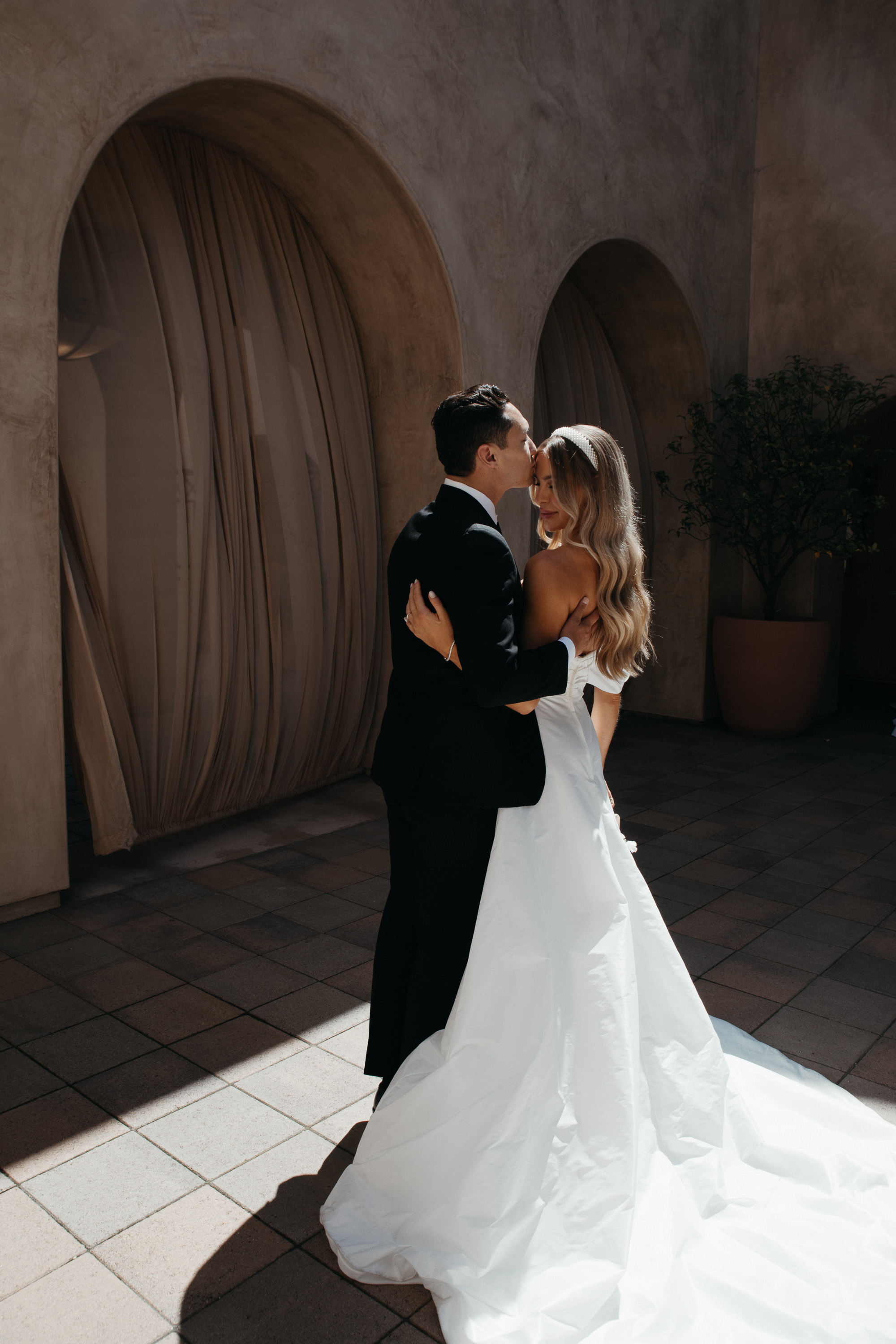 Groom in black suit kissing bride’s forehead as she stands in white wedding dress with long train under arches.