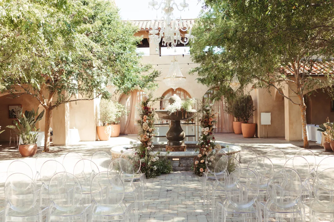 Outdoor wedding ceremony setup with transparent chairs facing a central fountain adorned with floral arrangements, under leafy trees.
