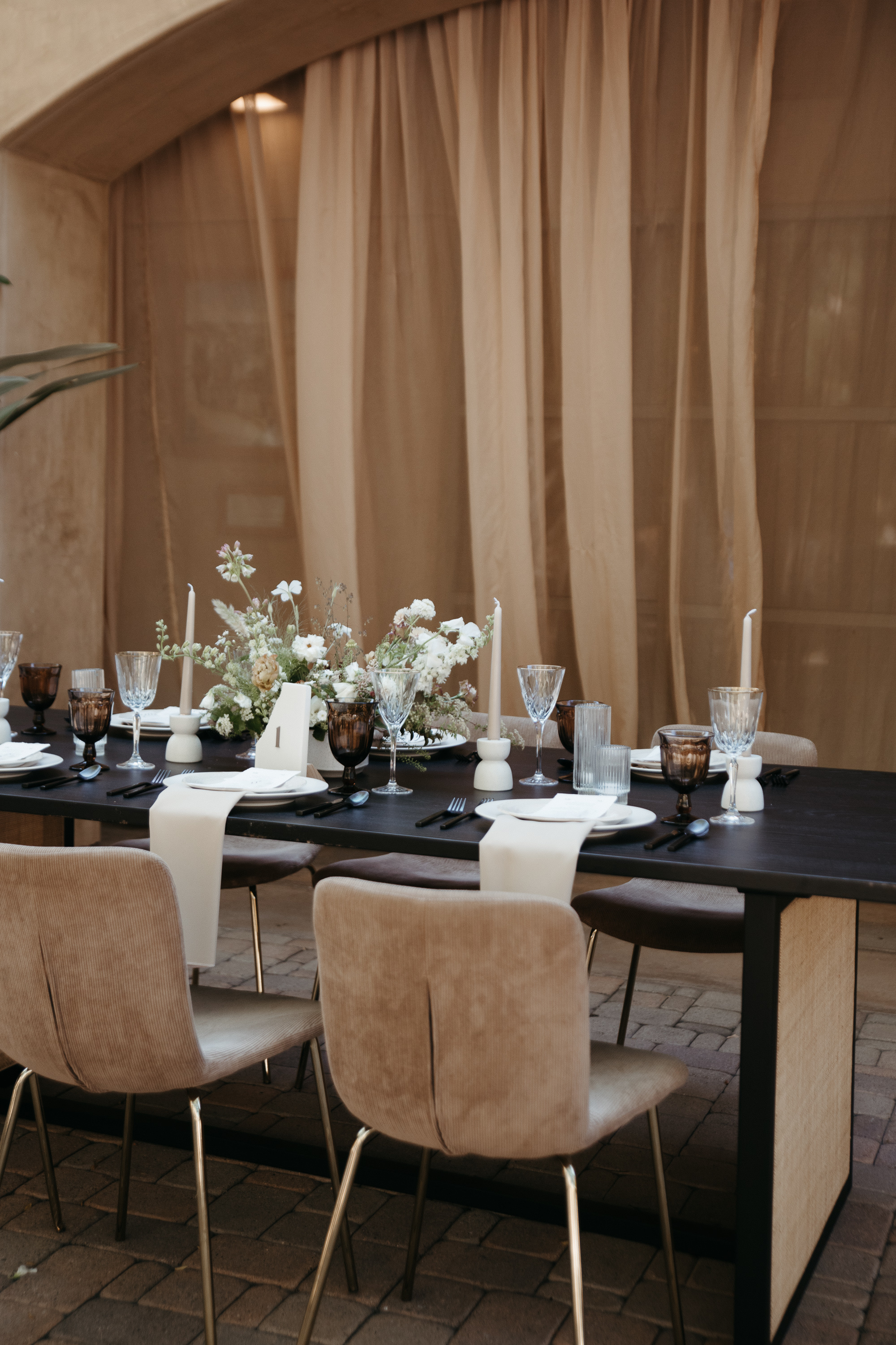 Elegant dining table set with white floral centerpiece, candles, and glassware, surrounded by beige cushioned chairs on a brick floor.