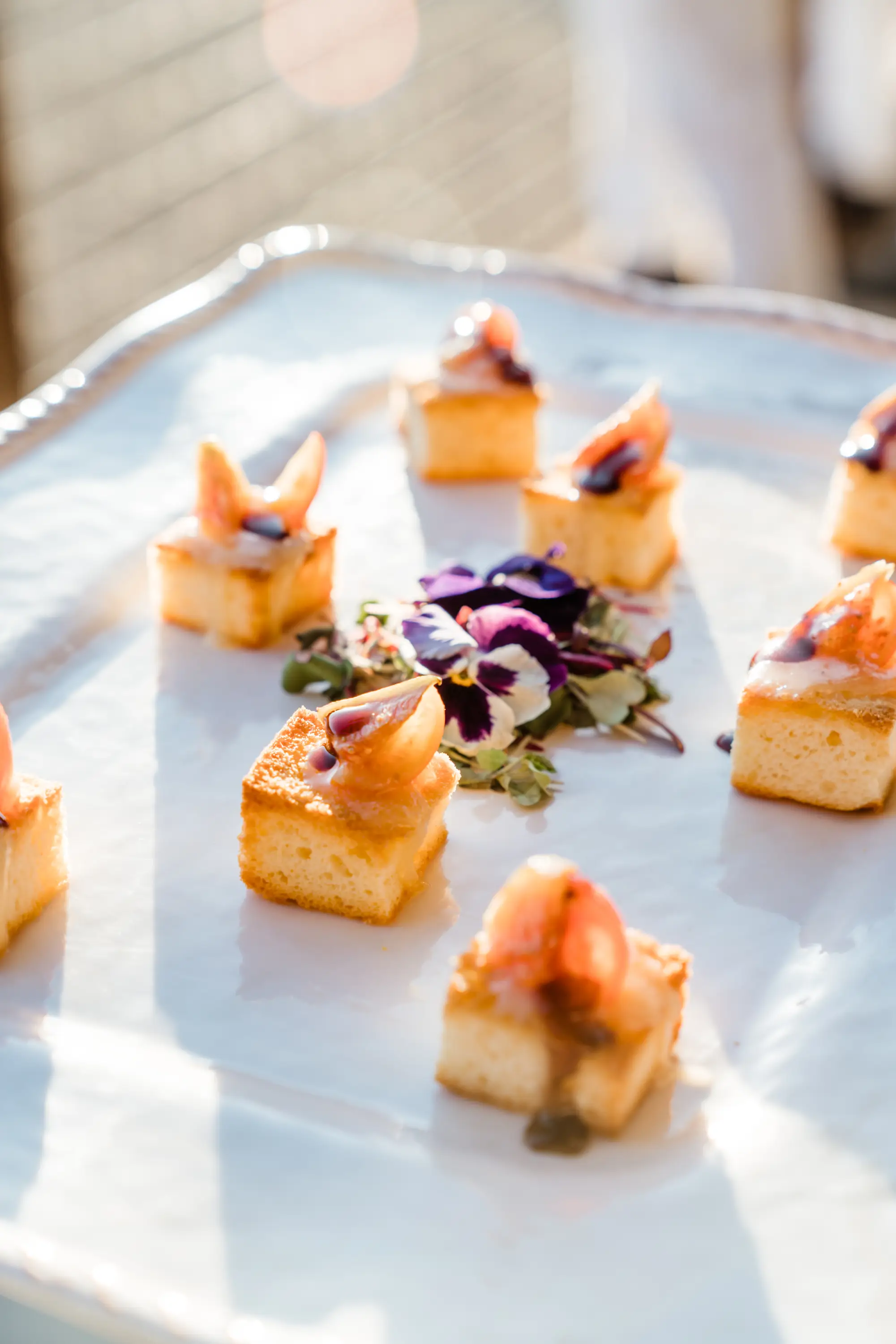 Small square pieces of toasted bread topped with fig slices, balsamic glaze, and microgreens arranged around edible flowers on a white platter.