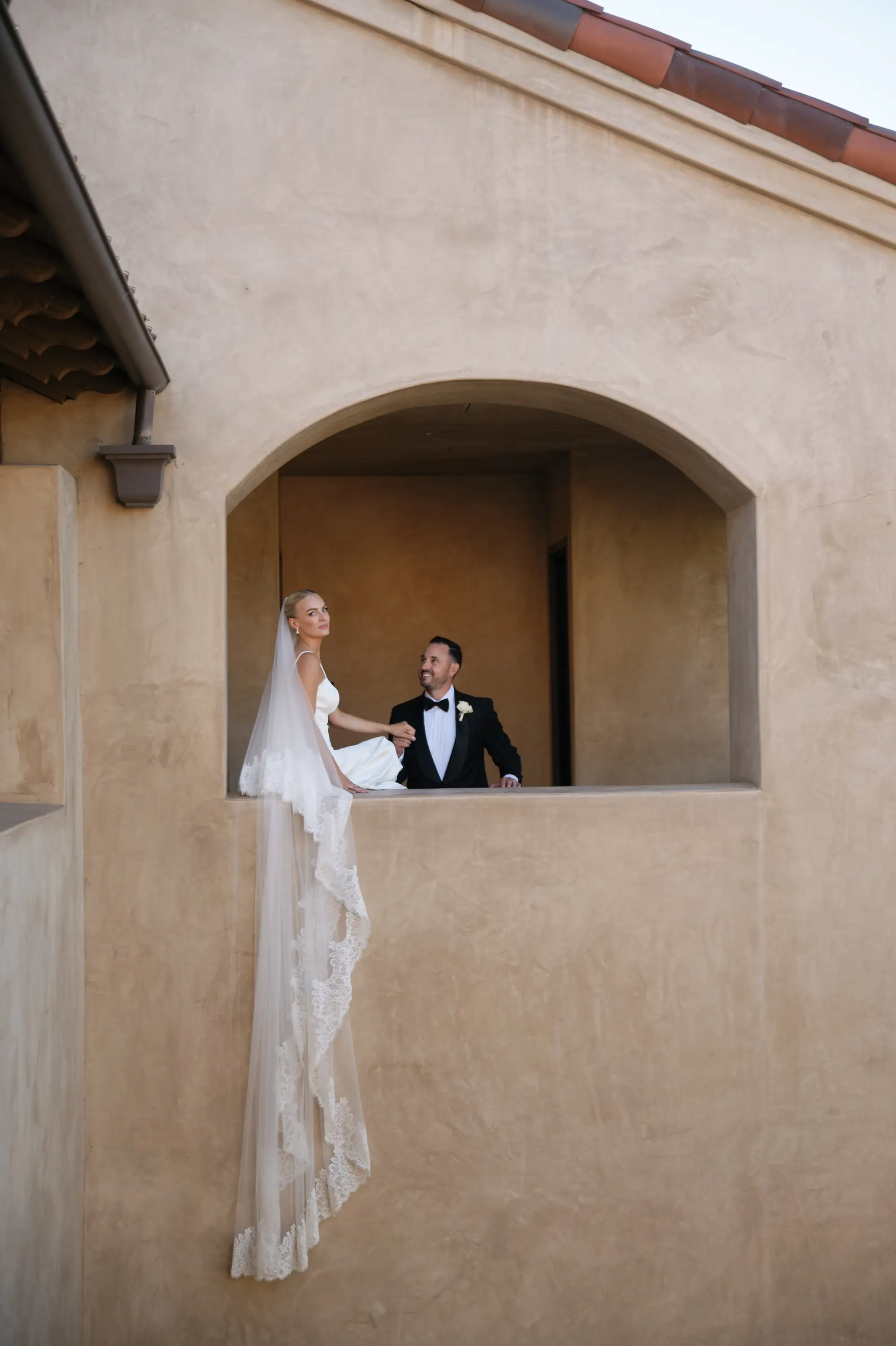 Bride in white dress and lace veil sitting on a ledge holding hands with a groom in a black tuxedo under an arched window in a beige stucco building.