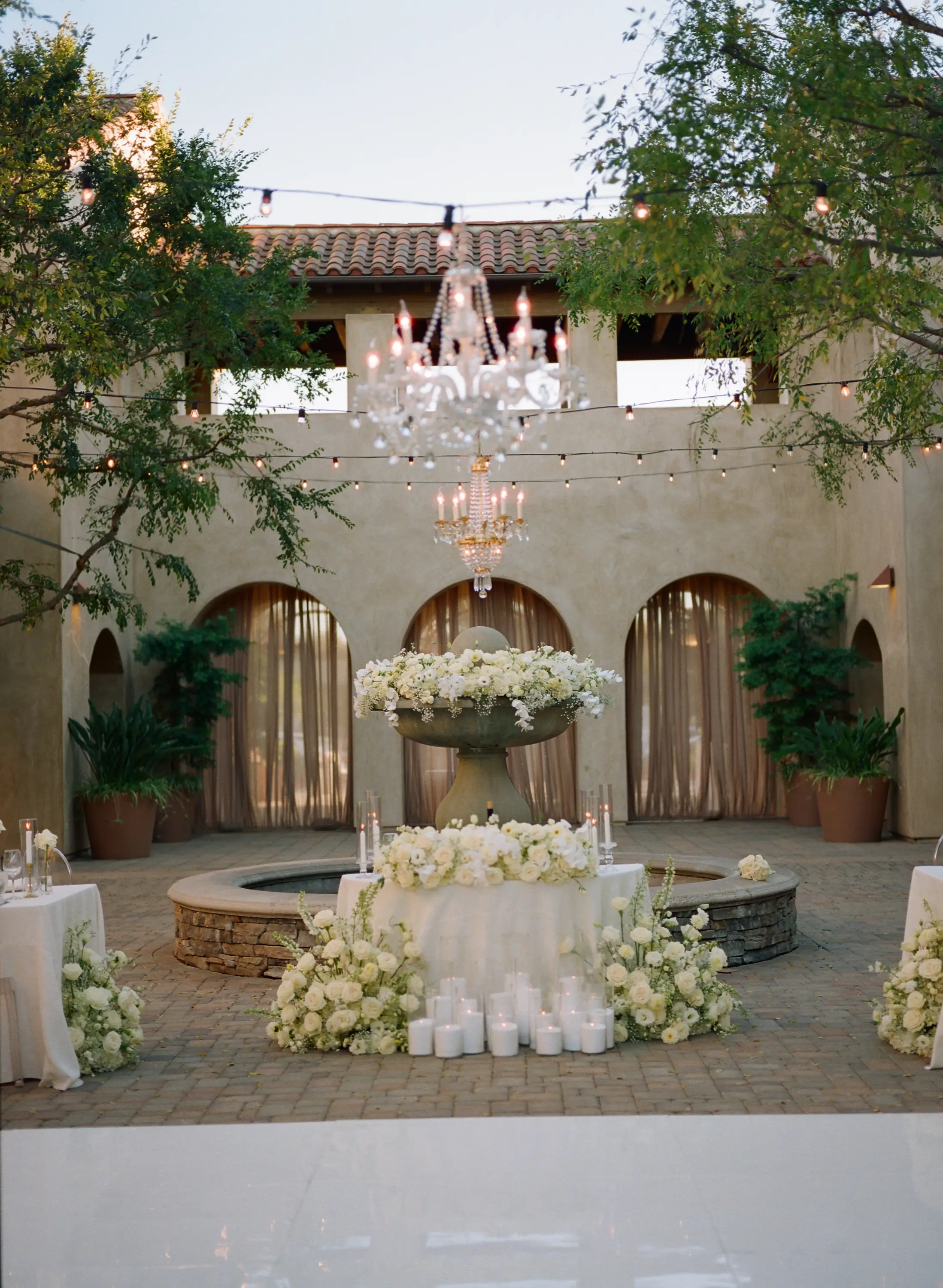 Outdoor wedding venue courtyard with a central stone fountain decorated with white flowers and candles, surrounded by potted plants and string lights overhead.