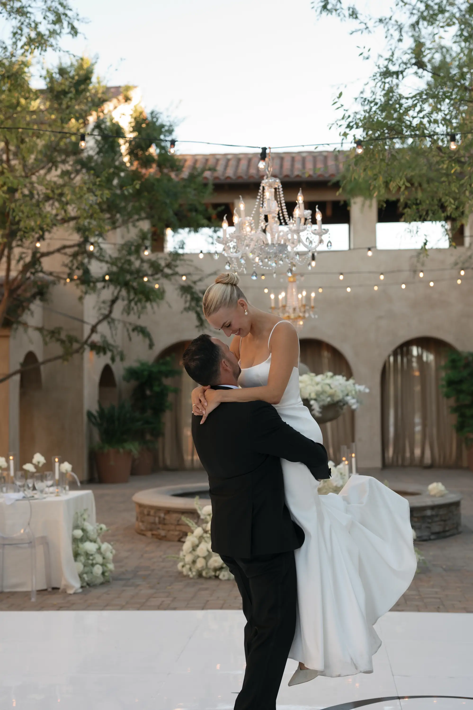 Groom lifting bride in white gown under chandeliers and string lights at outdoor wedding venue.