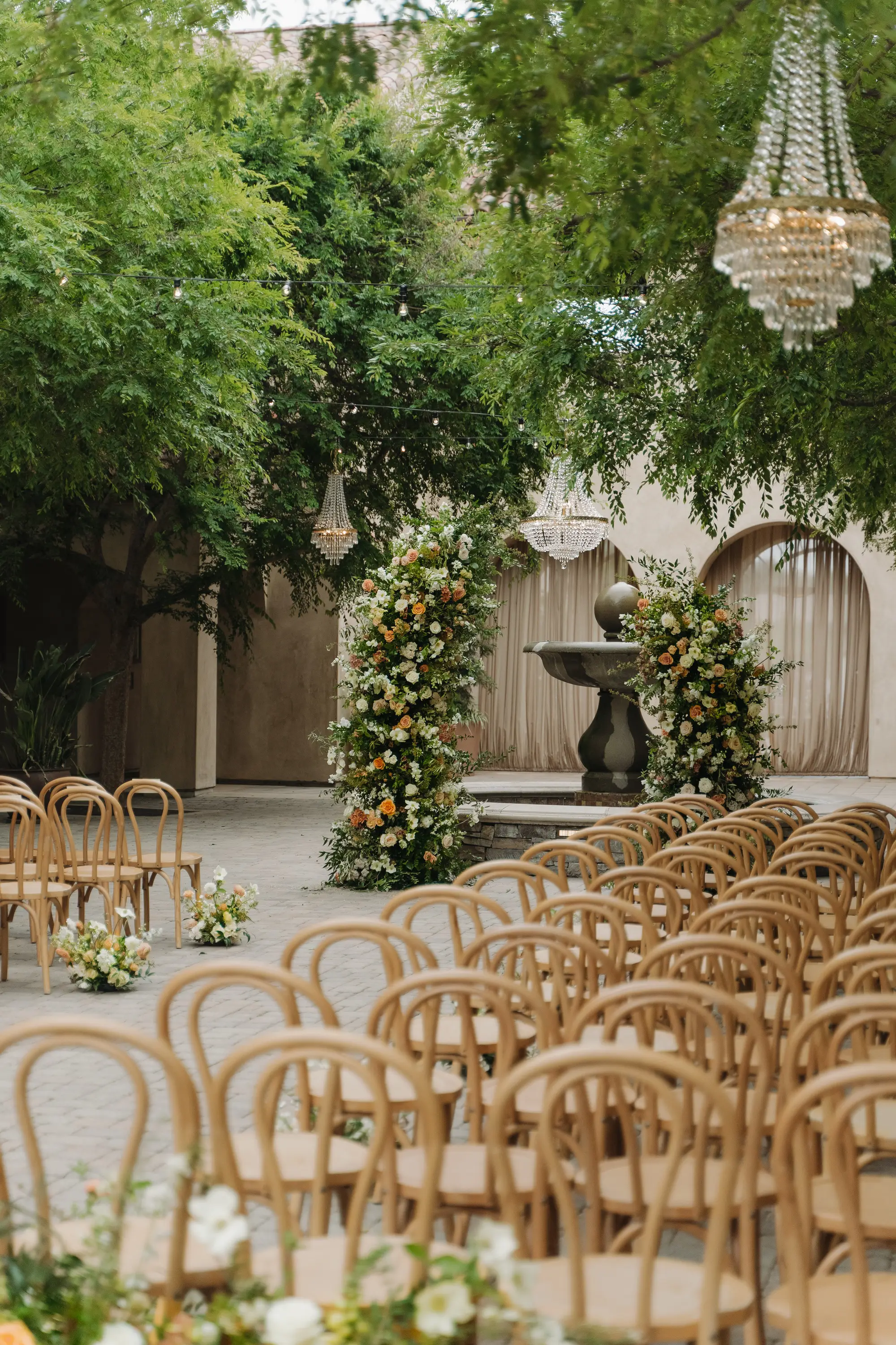 Outdoor wedding ceremony setup with wooden chairs facing floral archways and a stone fountain under green trees with hanging chandeliers.