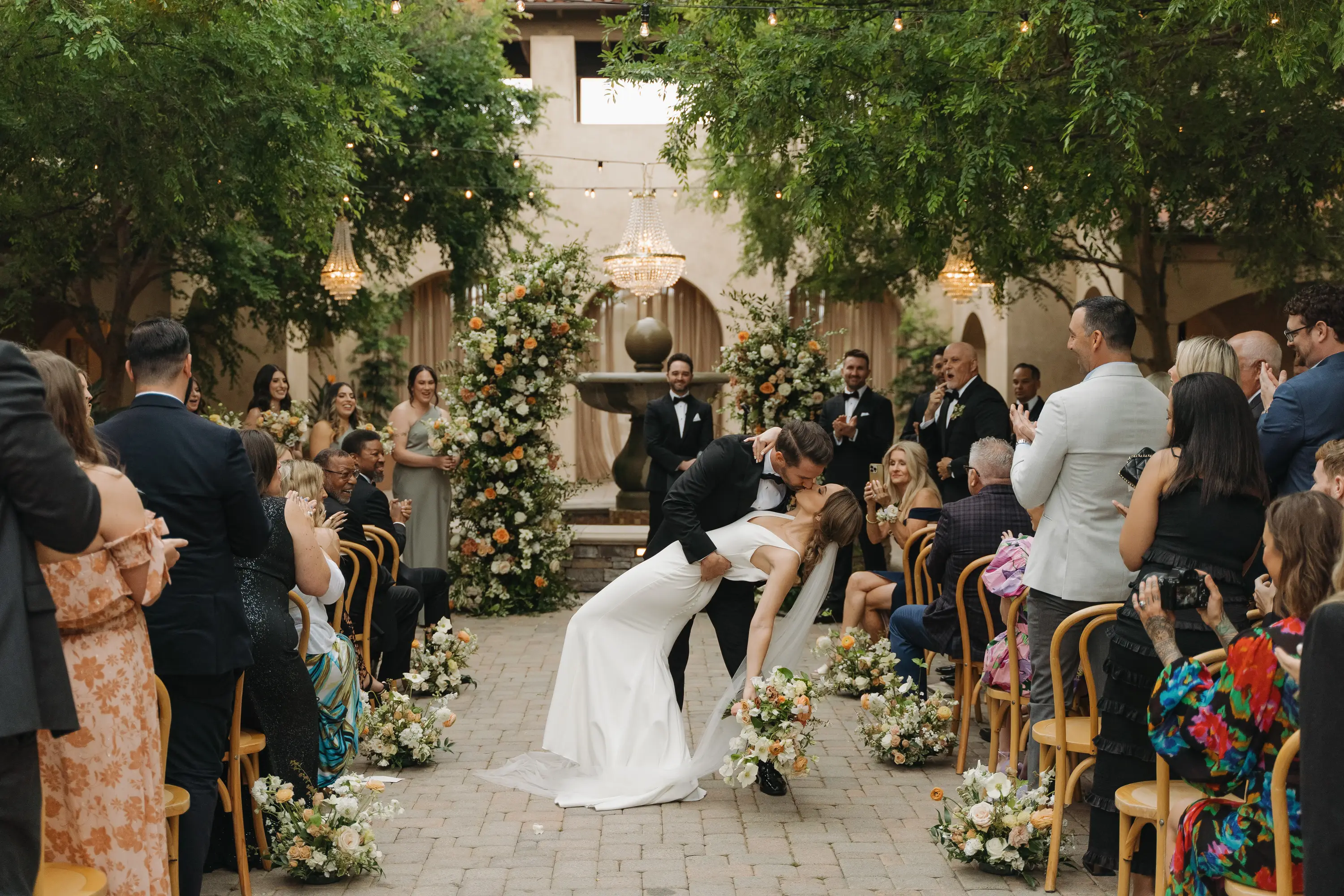 Bride and groom share a kiss while the groom dips the bride at an outdoor wedding ceremony with guests applauding.