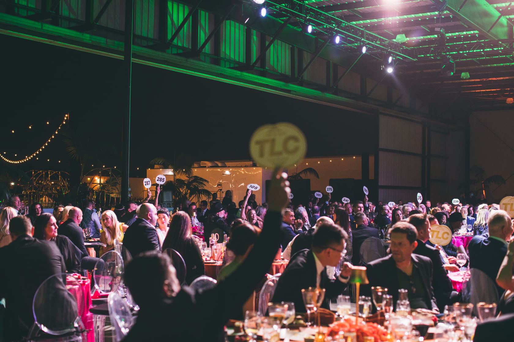People seated at round tables in a large indoor venue raising numbered paddles during an auction event.