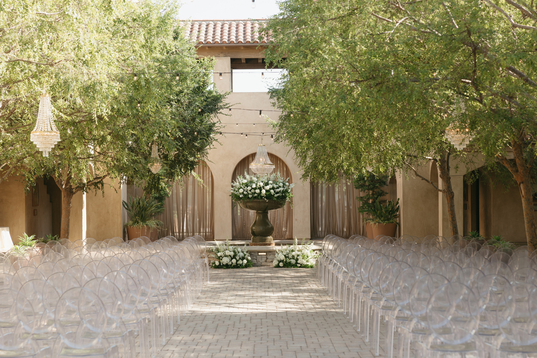 An outdoor ceremony with white flowers and greenery is set at Serra Plaza, a Spanish-style wedding venue located in San Juan Capistrano, California.