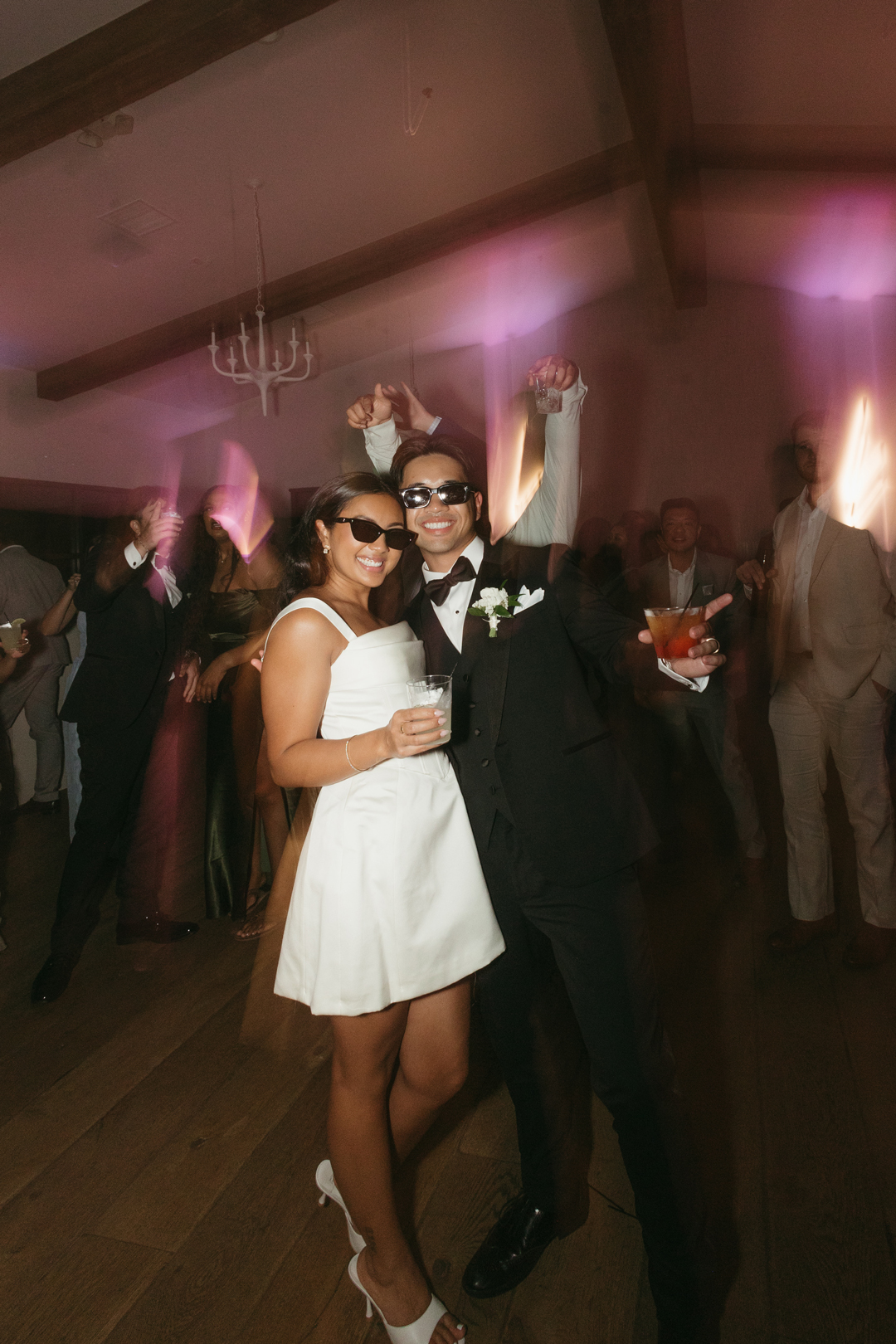 Bride and groom standing on dance floor with cocktails in hand wearing sunglasses.