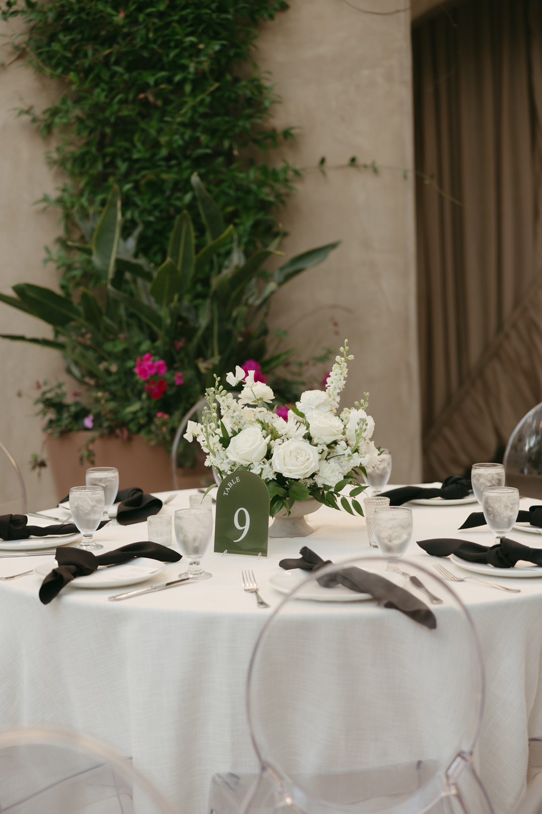 A white floral centerpiece sits at the center of a reception table. White plates and black linen napkins surround the table and a modern green table number sits next to the centerpiece.
