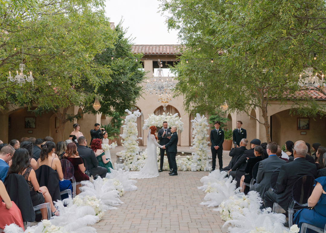 Guests sit on both sides of the aisle as the bride and groom recite their vows surrounded by white flowers.