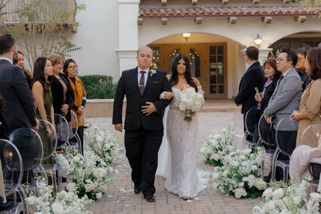 Bride and her father walk arm in arm down the aisle as guests watch from their seats.