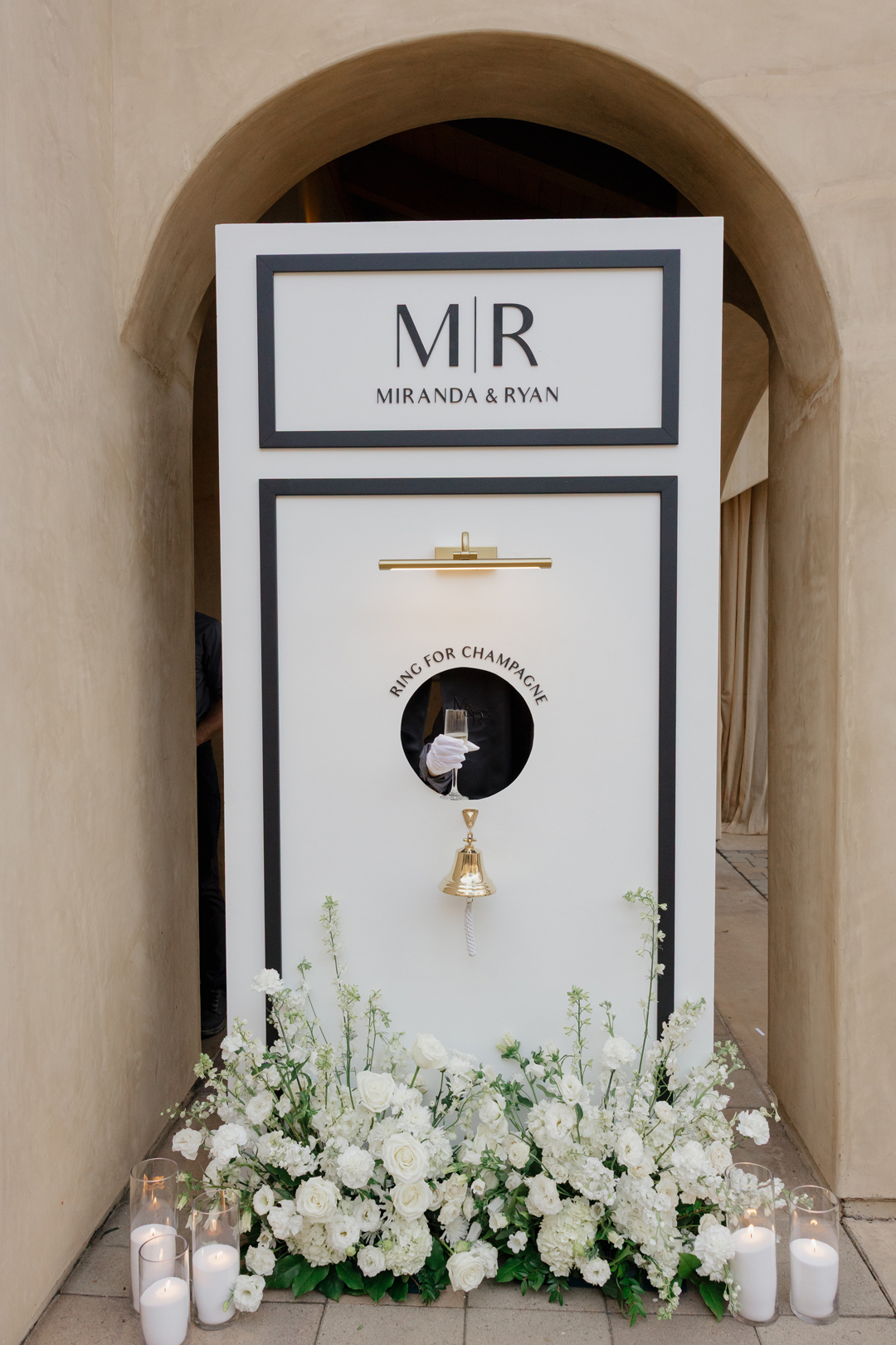 Arm with champagne in white-glove hand reaches out of 'ring for champagne' wall to greet guests with champagne as they arrive to the wedding venue.