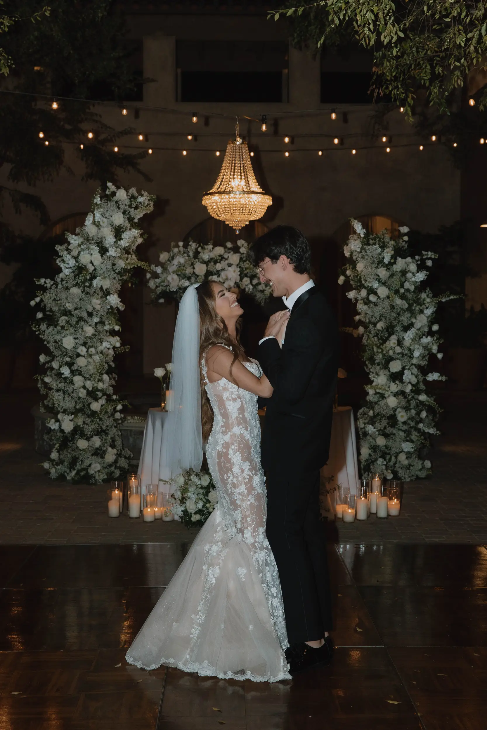 Bride and groom smile as they share their first dance on the wooden dance floor with their white floral sweetheart table as their backdrop.