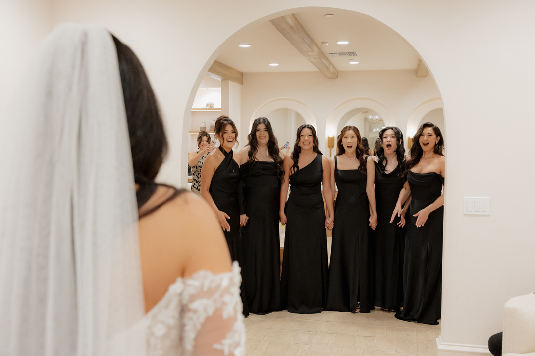 Bridesmaids dressed in black are happily surprised when they see the bride in her lace wedding dress inside the Serra Plaza Spanish Revival wedding venue Bridal Suite.