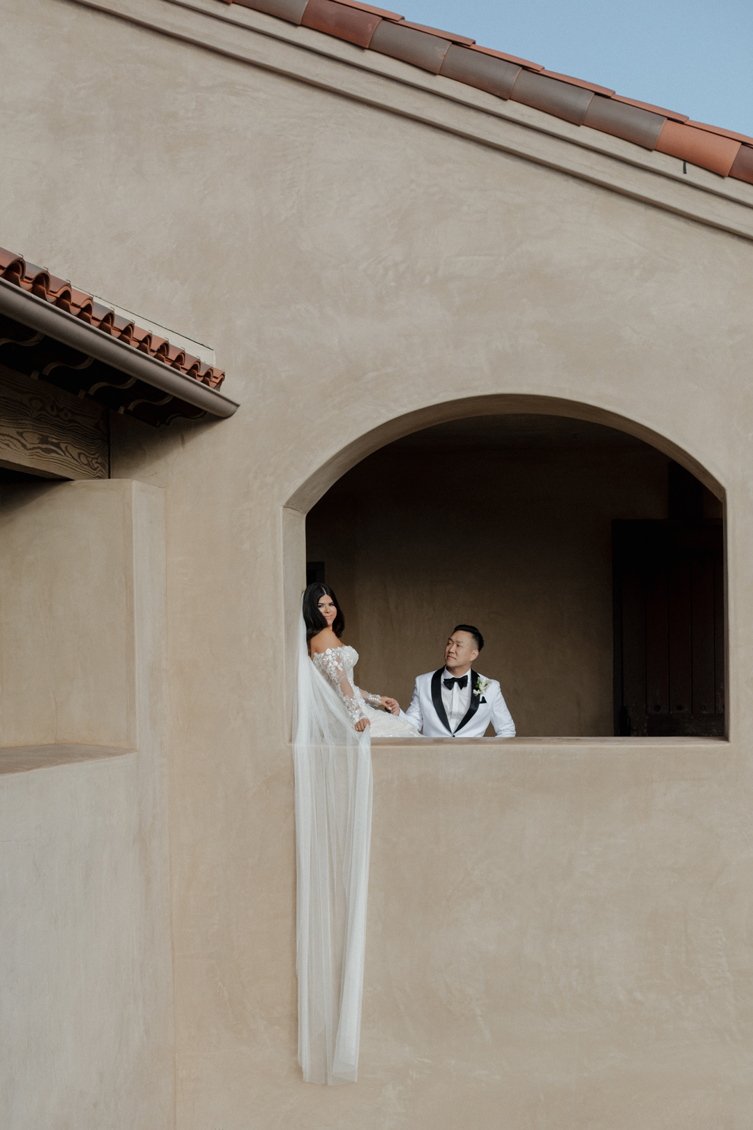 Bride and groom pose in open window frame on the second story with bride's veil cascading down the venue walls.