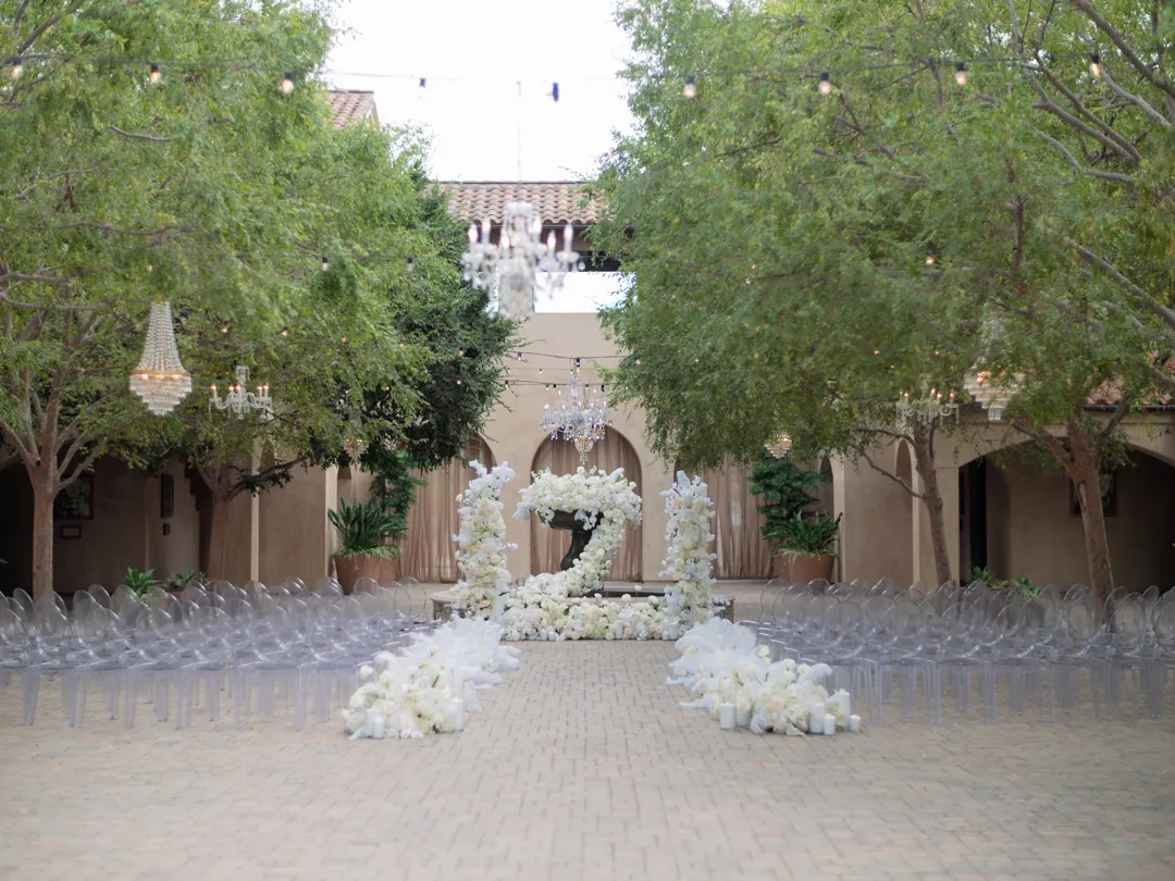 White flowers and feathers, sparkling chandeliers, and a spanish-style wedding venue backdrop complete the scene for this courtyard wedding ceremony.