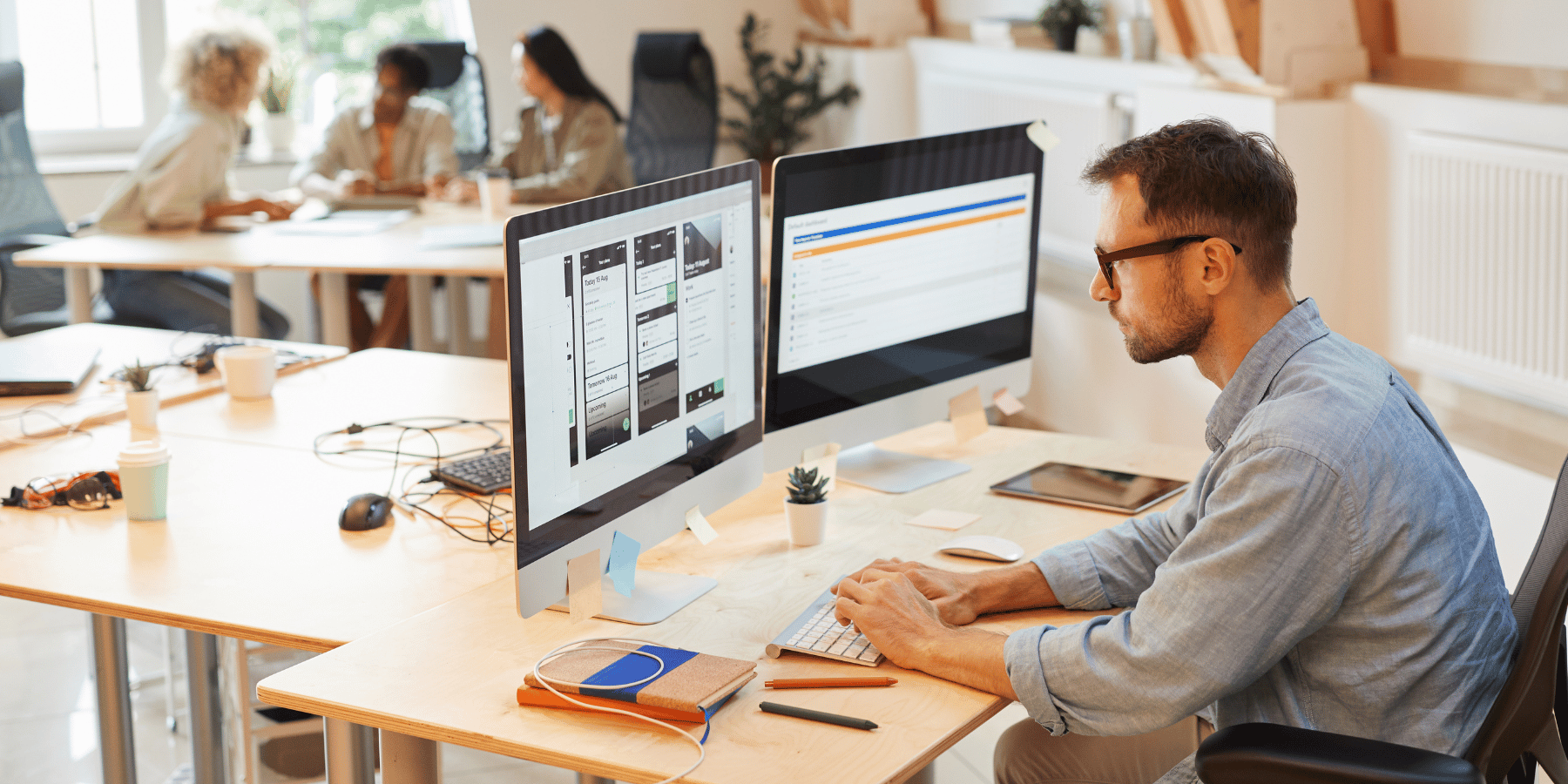 Mann arbeitet konzentriert am Computer in einem modernen Büro mit Kolleginnen im Hintergrund.