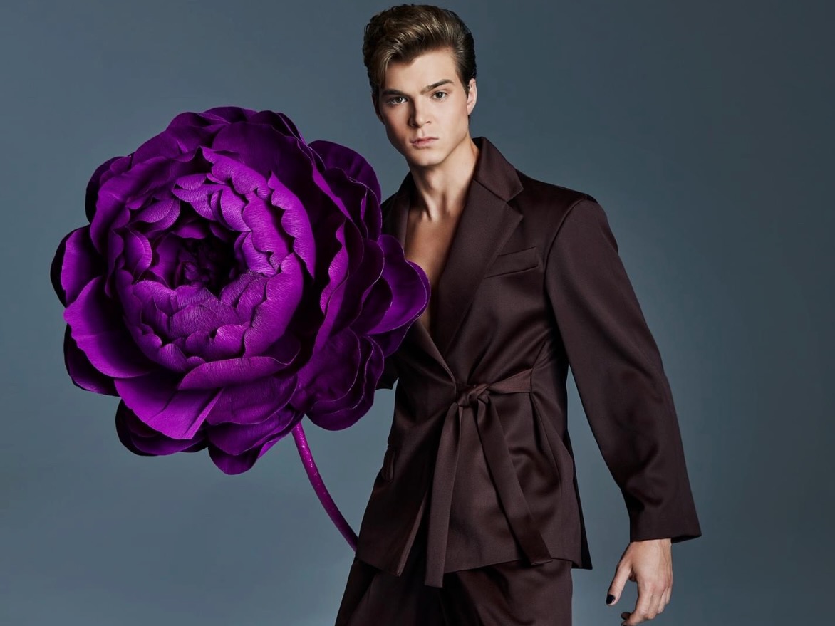 Sesja zdjęciowa Top Model: mężczyzna w garniturze trzymający wielkoformatowy fioletowy kwiat Fiori di Matis