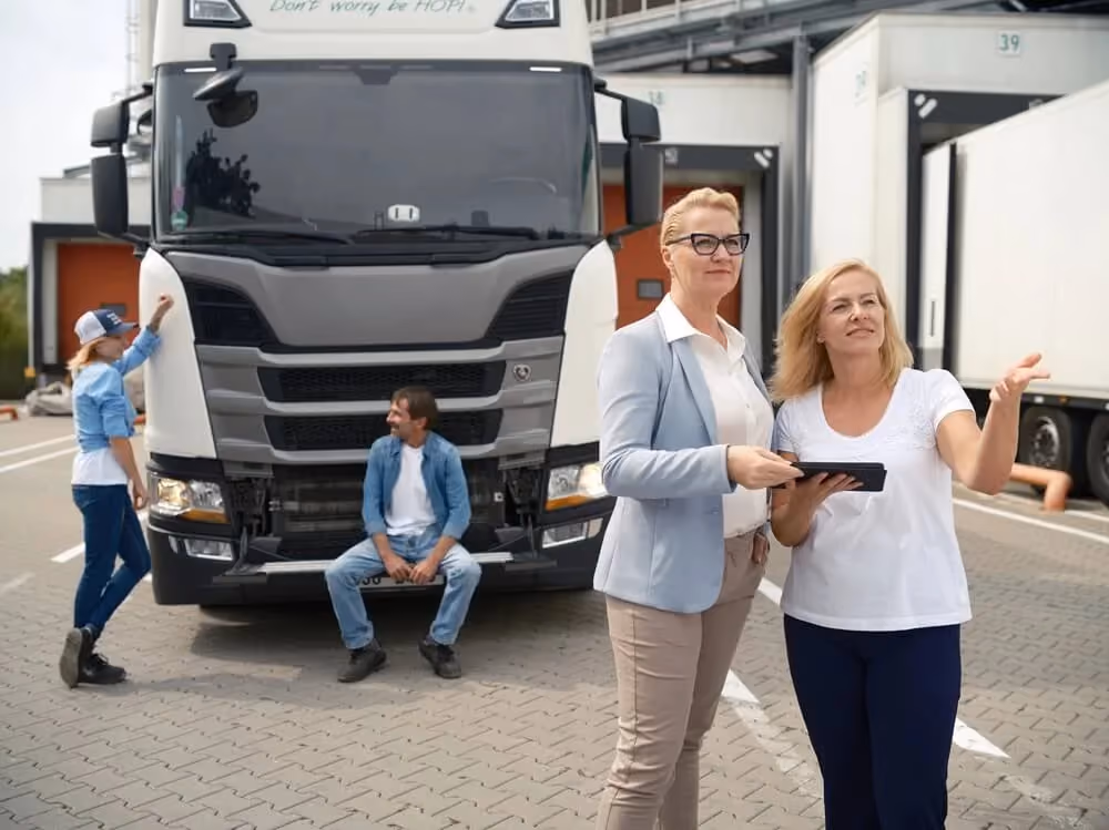 Two women discussing near two people by a parked large white truck in a loading dock area.