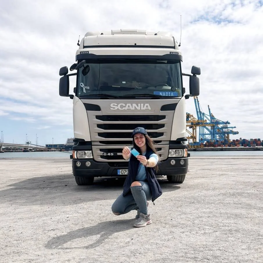 Woman kneeling in front of a white Scania truck in an industrial port holding a blue card.