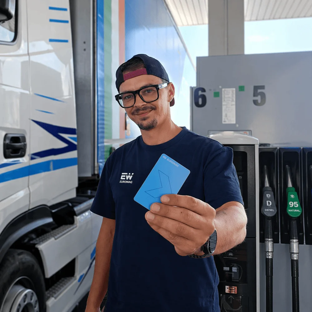Man in glasses and hat at gas station holding a blue card near fuel pumps and white truck.