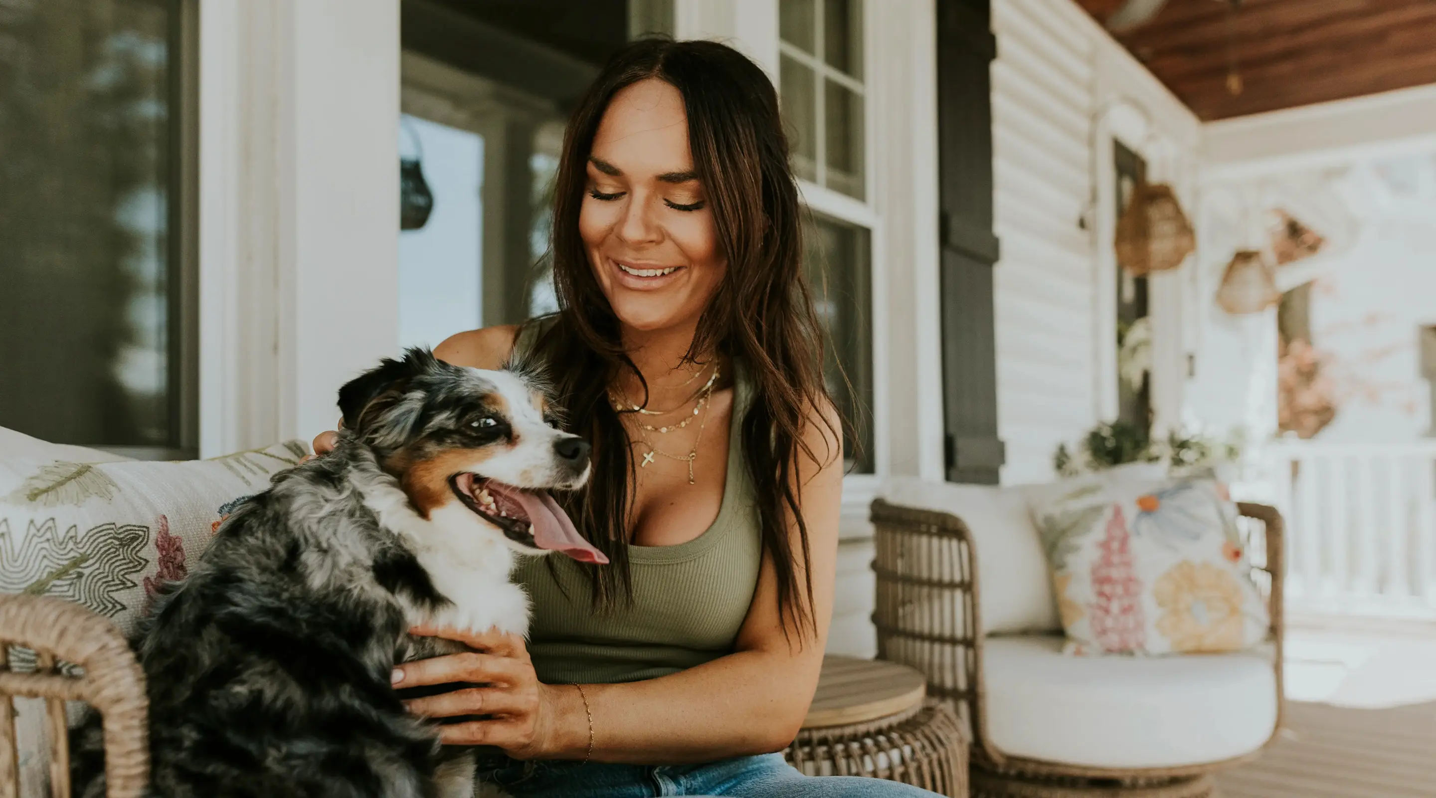 Smiling woman with long dark hair sitting on a porch and petting a happy Australian Shepherd dog.