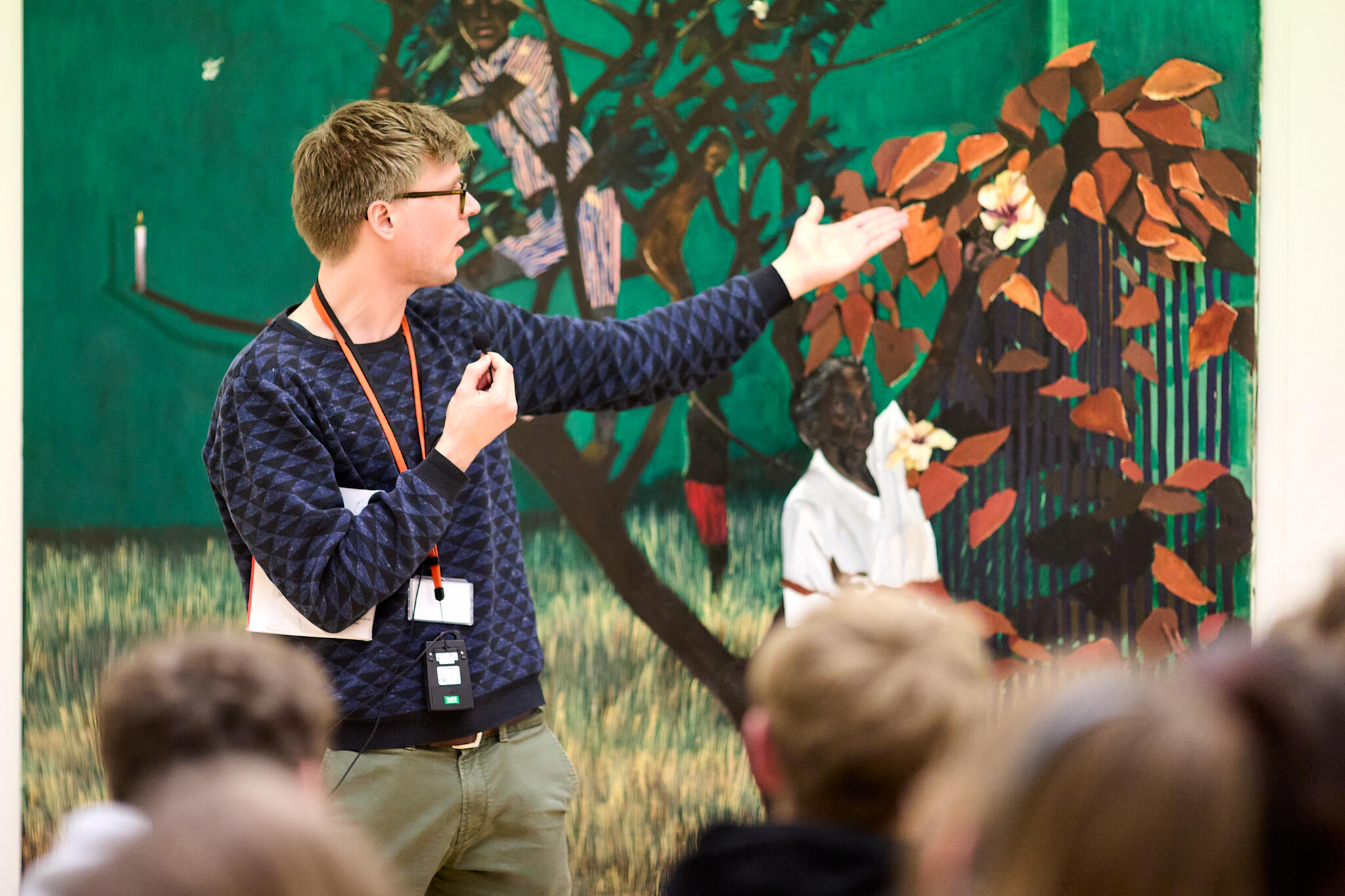 Een mannelijke museumdocent staat voor een groen schilderij. Hij wijst naar rechts en praat. Op de voorgrond zie je de achterhoofden van luisterende studenten.