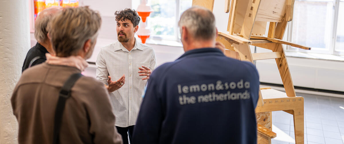 Een jonge kunstenaar staat bij een kunstwerk van op elkaar gestapelde houten stoelen. Hij spreekt een groep bezoekers toe.