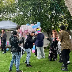 Group of people outdoors holding pink and blue balloons, with one person holding a birthday cake under a pink cloth.