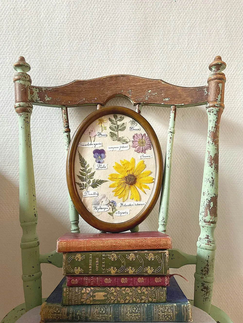 Chaise vintage en bois patiné accueillant un cadre floral ovale posé sur une pile de livres anciens.