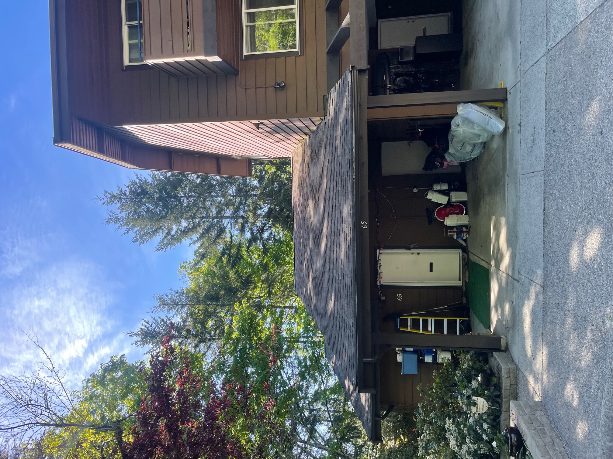 A residential building featuring a brown exterior and a carport. The carport contains various items, and trees are visible in the surrounding area. The sky is clear with some clouds, typical of a sunny day.