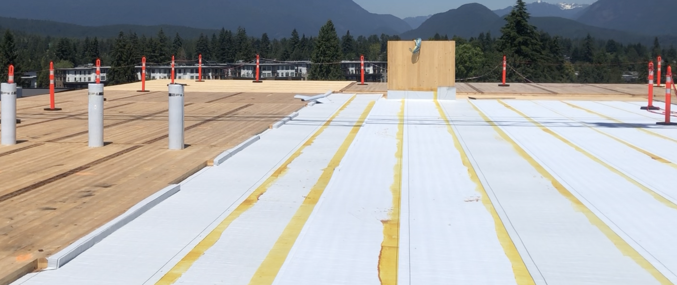 A flat commercial roofing surface under construction, featuring white insulation material and yellow lines. Red safety cones are placed around the perimeter, with mountains visible in the background.