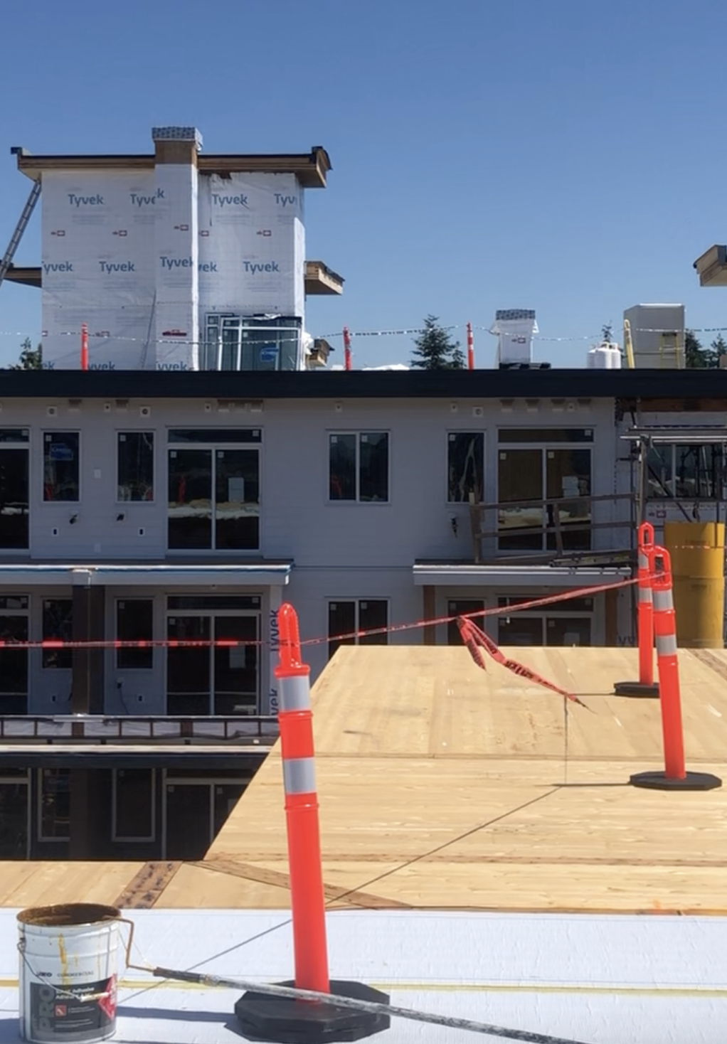 A construction site featuring a wooden roof installation with safety cones and caution tape. Nearby, a building is undergoing exterior work, covered in Tyvek, with windows and scaffolding visible.