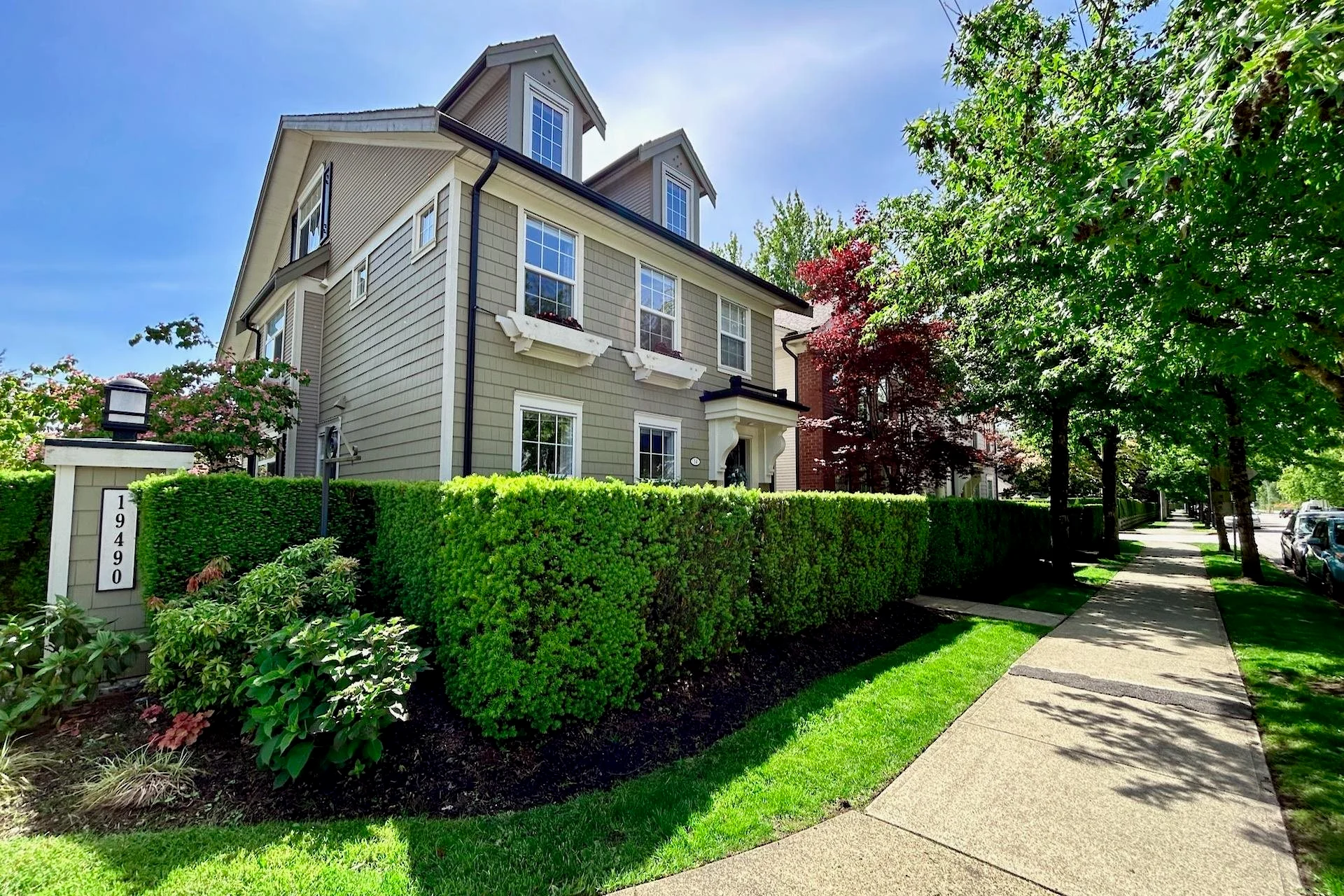 A two-story grey house in Pitt Meadows, surrounded by neatly trimmed hedges and colorful plants. A wide sidewalk runs alongside the front yard, and tall trees provide shade in the area.