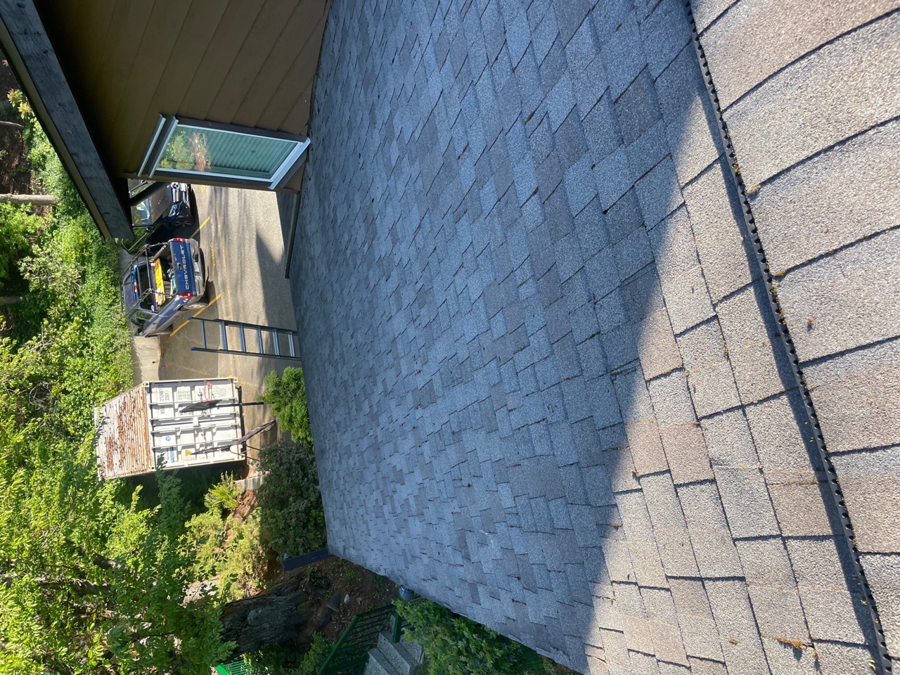 A sloped roof covered with gray asphalt shingles, with a view of a nearby wooden building and some greenery in the background. A ladder leans against the building, and a parked vehicle is visible in the driveway.