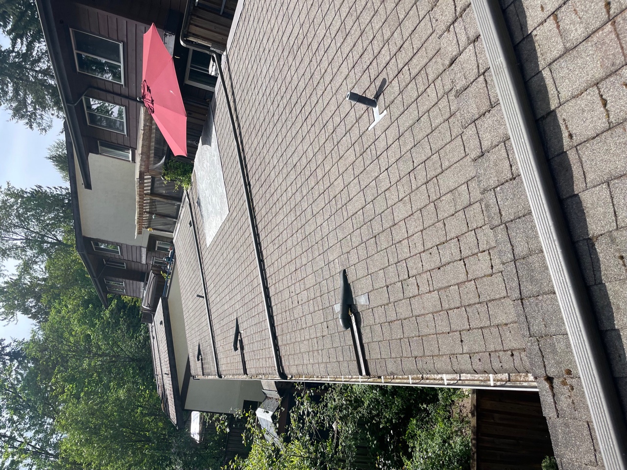 A flat rooftop of a house covered with gray shingles, with a red umbrella visible on a deck nearby. Surrounding trees and part of the house's siding can be seen in the background.