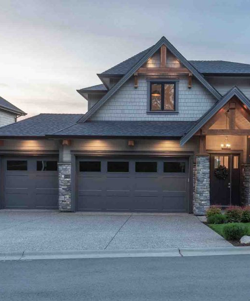 A modern house featuring a two-car garage with dark doors, accented by stone and wood elements. Soft lighting highlights the architectural details under a pastel sky, representing a Langley house roof installation.
