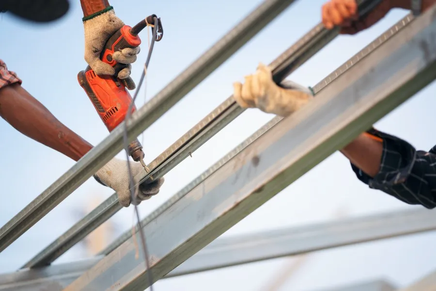 Two hands working on a construction project, one holding a power tool and the other guiding a metal pole. The setting appears to be outdoors, with a clear blue sky in the background.