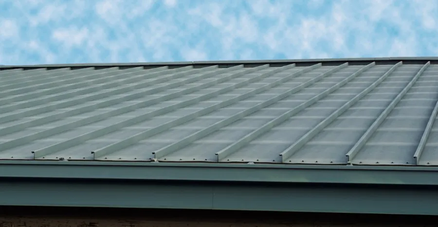 A close-up view of a light-colored metal roof, showcasing its ridged and textured surface against a cloudy blue sky.