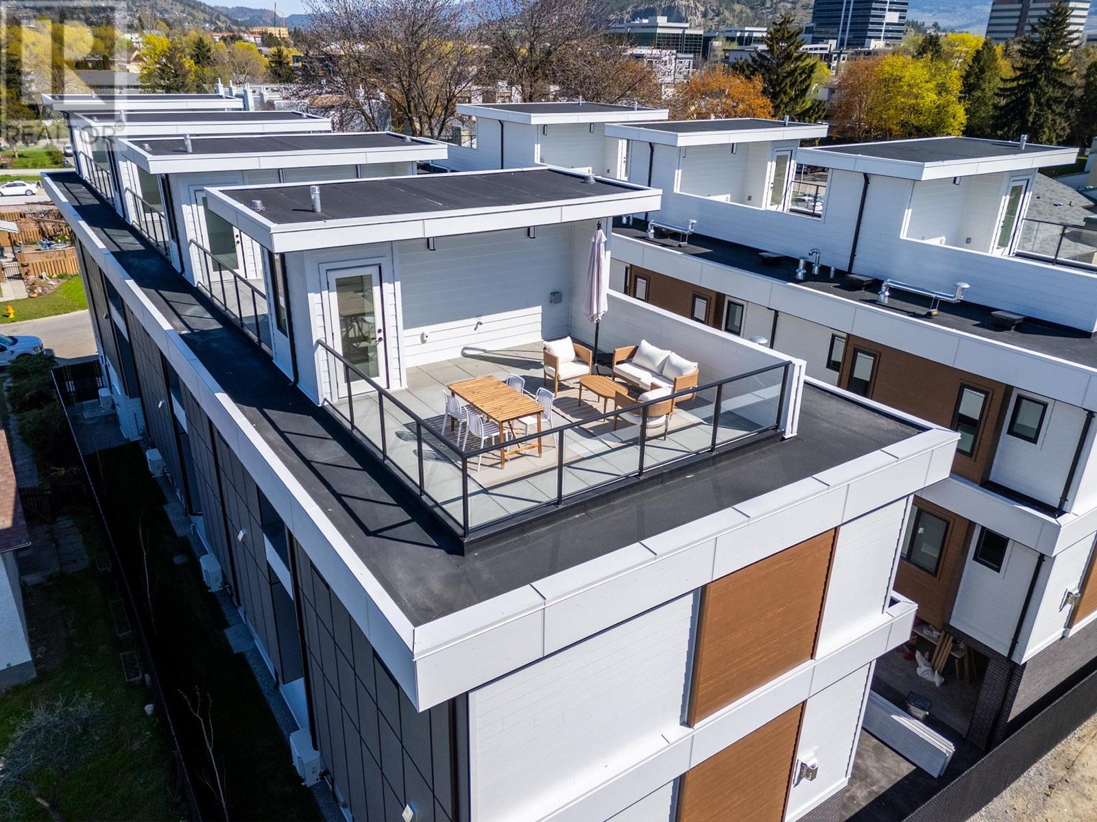 Aerial view of a modern white condo building with a rooftop terrace in Kelowna. The terrace features a seating area with chairs and a table, surrounded by glass railings. The surrounding landscape includes trees and distant buildings.