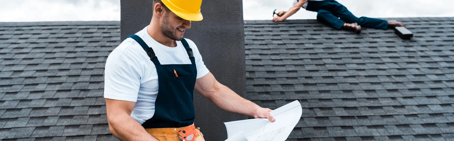 A construction worker wearing a yellow hard hat and overalls examines blueprints on a rooftop, while another worker is seen installing shingles in the background. The image captures a sunny day with scattered clouds, emphasizing the home shingle roof installation process.