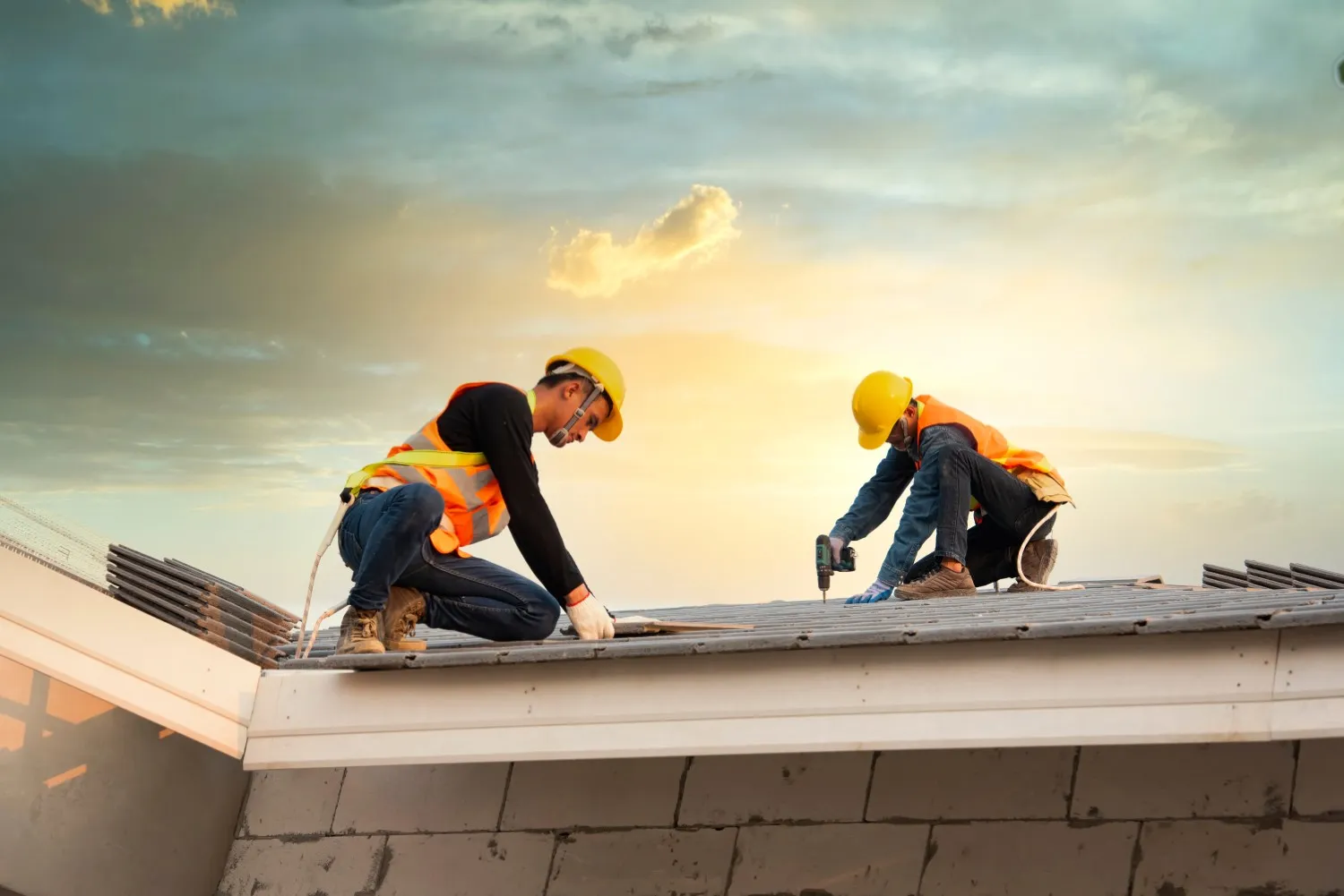 Two construction workers wearing yellow helmets and orange vests are installing roofing materials on a residential building. The sun sets in the background, casting a warm glow over the scene, emphasizing the importance of commercial roofing work.