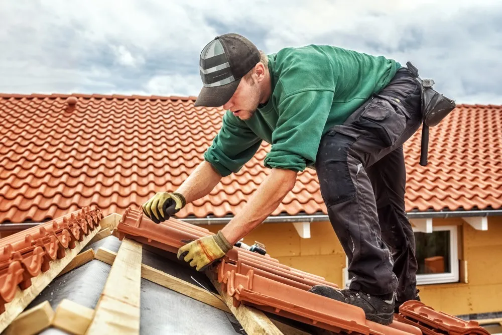 A person installing roof tiles on a sloped roof, wearing gloves and a cap. The roof features red tiles, and the background includes a building with light-colored walls.