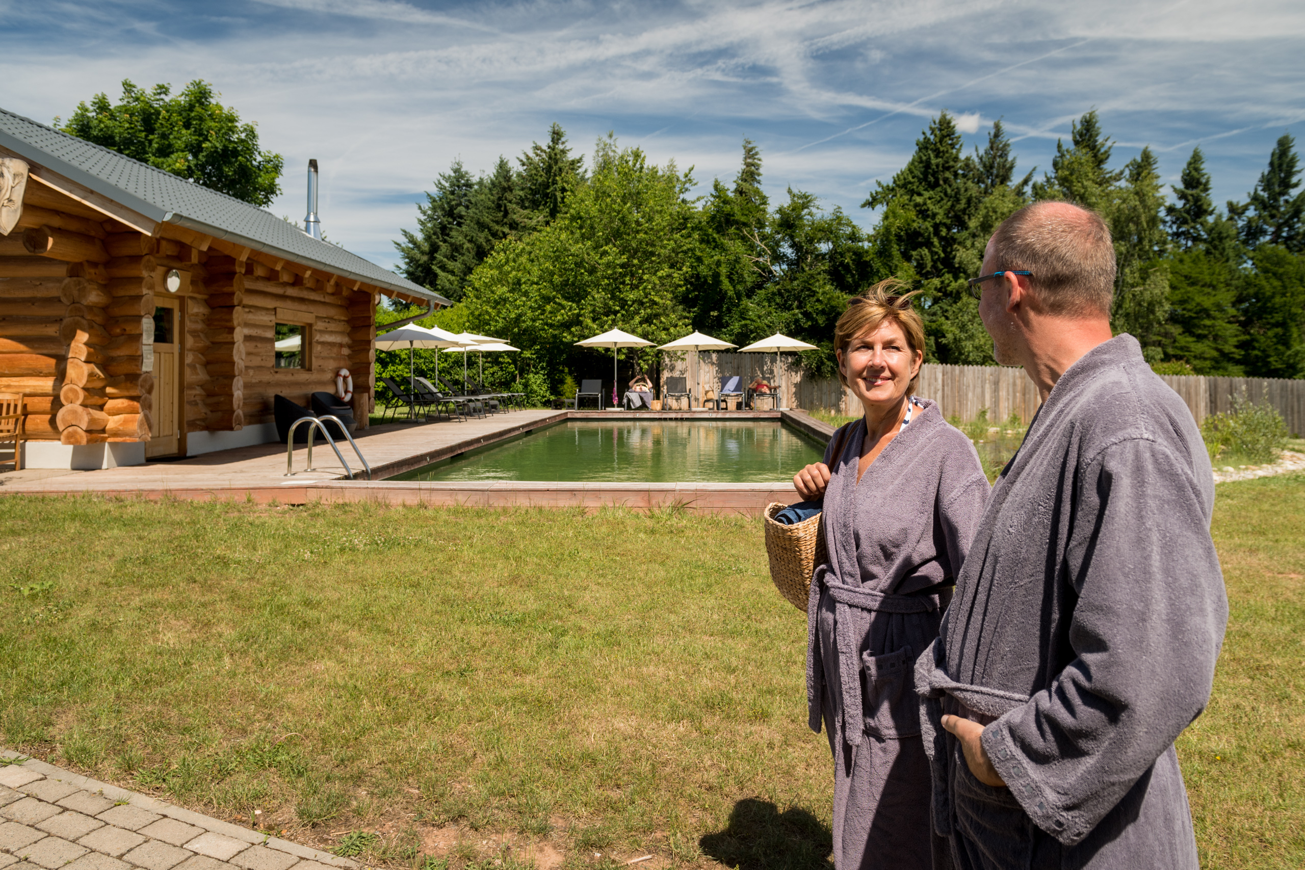 Zwei Personen in Bademänteln neben einem Naturpool mit Holzhütte